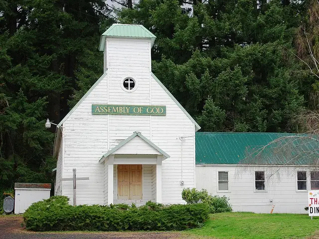 Small blue clapboard Assembly of God church in Buxton, Oregon, with a white cross on the gable and an American flag by the front steps.
