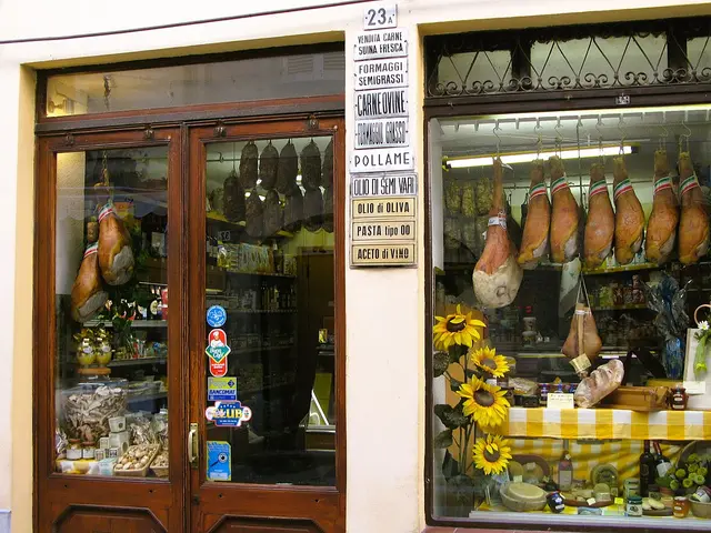 An Italian delicatessen storefront window with whole prosciutto legs and cheese wheels hanging on display, alongside sunflowers and olive oil bottles.