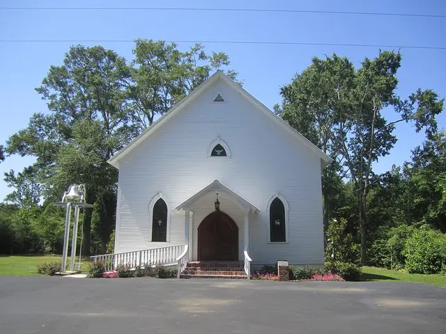 Small white wooden Christian church with a covered entrance porch and a freestanding bell, surrounded by trees