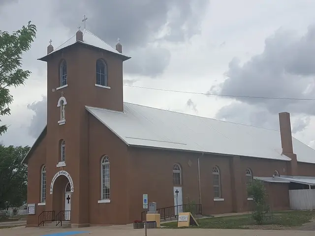 Adobe-style Catholic church with a tall bell tower topped by three crosses, under a cloudy sky