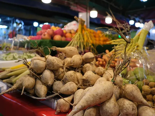 Fresh root vegetables piled high at an outdoor market produce stall, with more fruit and produce visible in the background.
