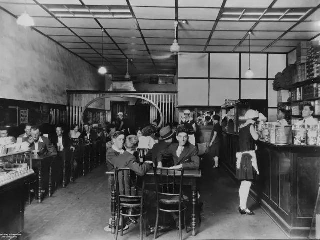 A 1929 photograph of a cafe interior, with the owner behind the counter and a waitress serving a customer.