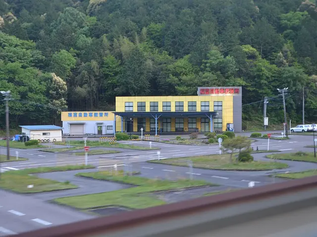 A yellow driving school building beside a car test course with road markings, set in a wooded valley in Japan.