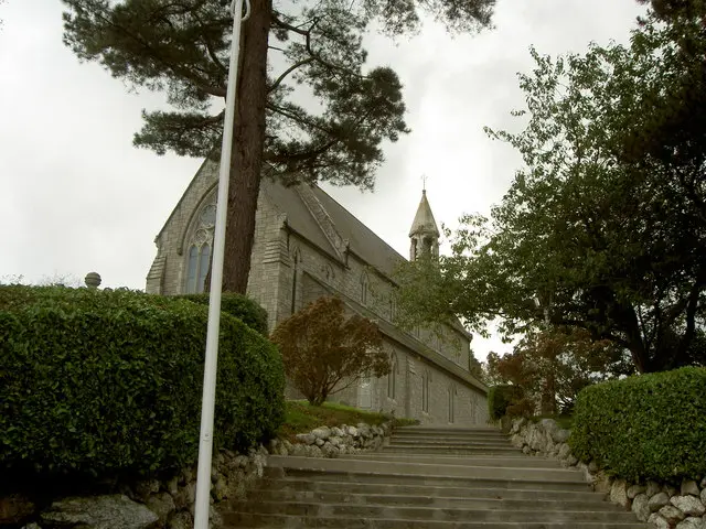 Exterior of Rathdrum Roman Catholic Church, a grey stone church with a small spire, seen through trees and hedges.