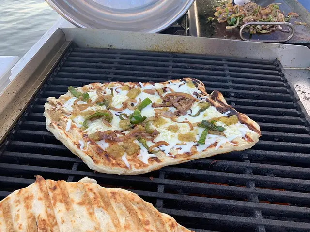 A small rectangular pizza being grilled outdoors over a gas flame.