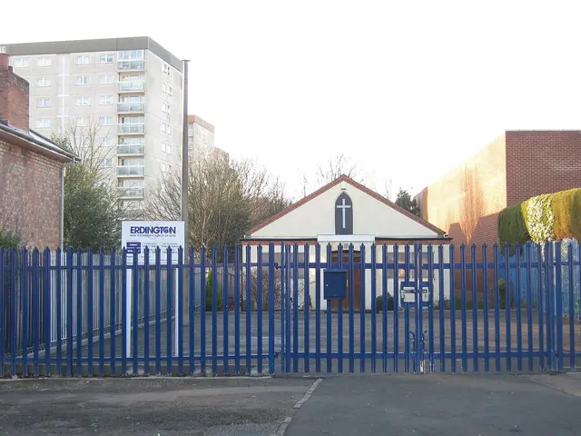 Exterior of a small place of worship building with a plain cross above the entrance, in Erdington, England, seen behind a security fence.