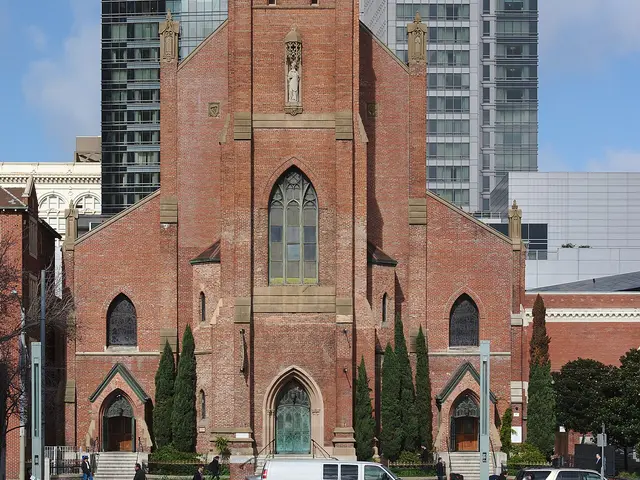 Red brick Gothic Revival Catholic church facade with a twin-lancet bell tower, dwarfed by modern glass skyscrapers behind it