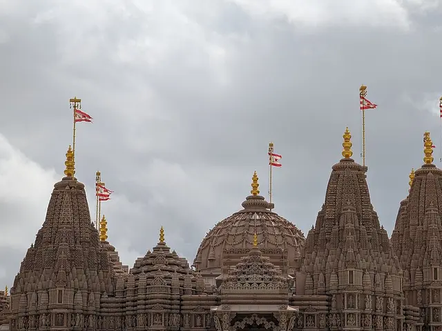 Carved sandstone-coloured shikhara towers of a Hindu mandir topped with flags, seen against a cloudy sky.