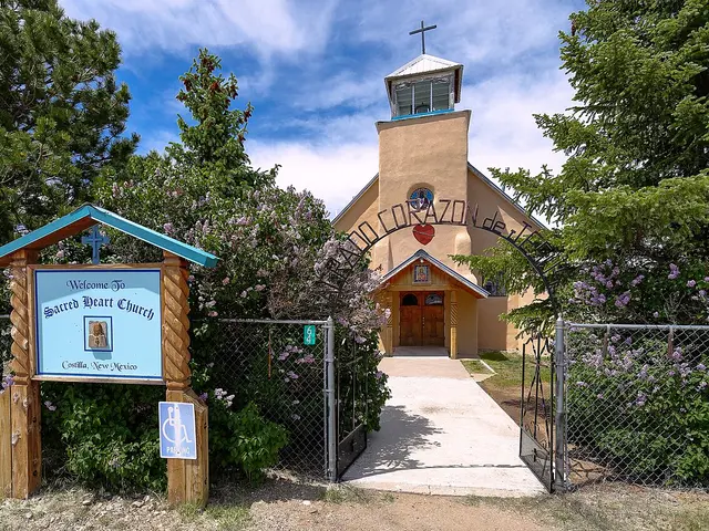 Small adobe-style Catholic church named Sacred Heart, seen through a garden gate framed by lilac bushes