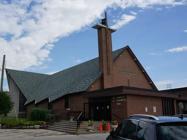 Modern red brick Catholic church with an angular roofline and a tall cross-topped spire, parking lot in foreground