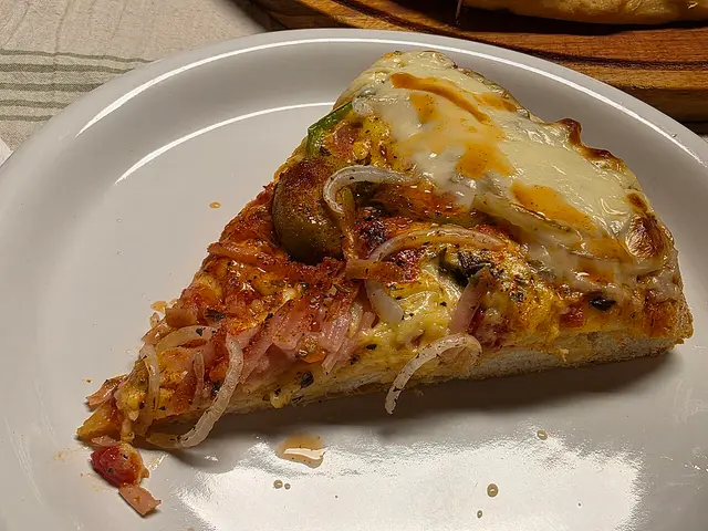 A close-up of a slice of Argentine-style pizza topped with melted cheese, ham, onion, and a green olive.